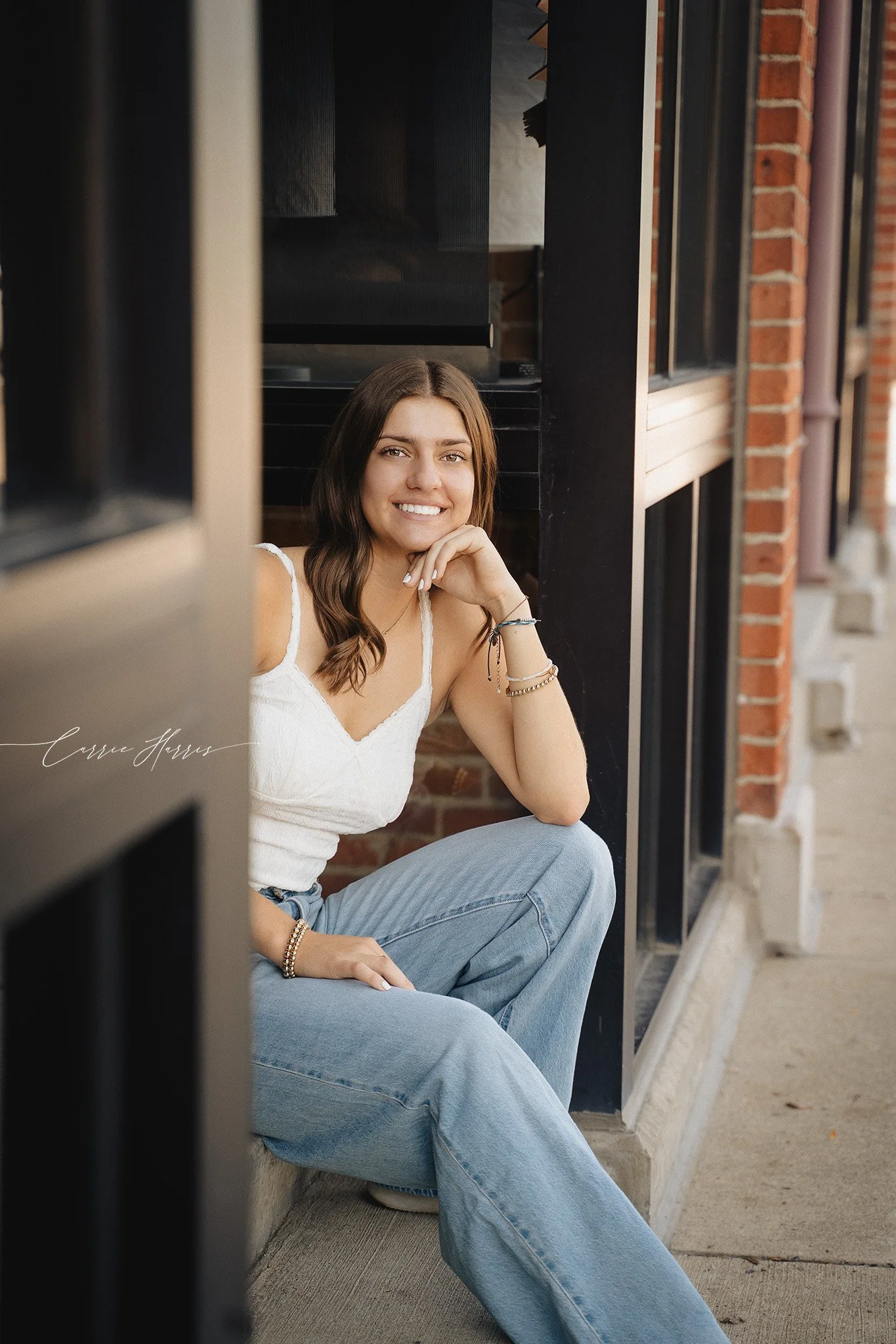 A young woman with long brown hair, smiling, sitting against a brick wall near a black window frame, wearing a white top and blue jeans, with jewelry on her wrist.