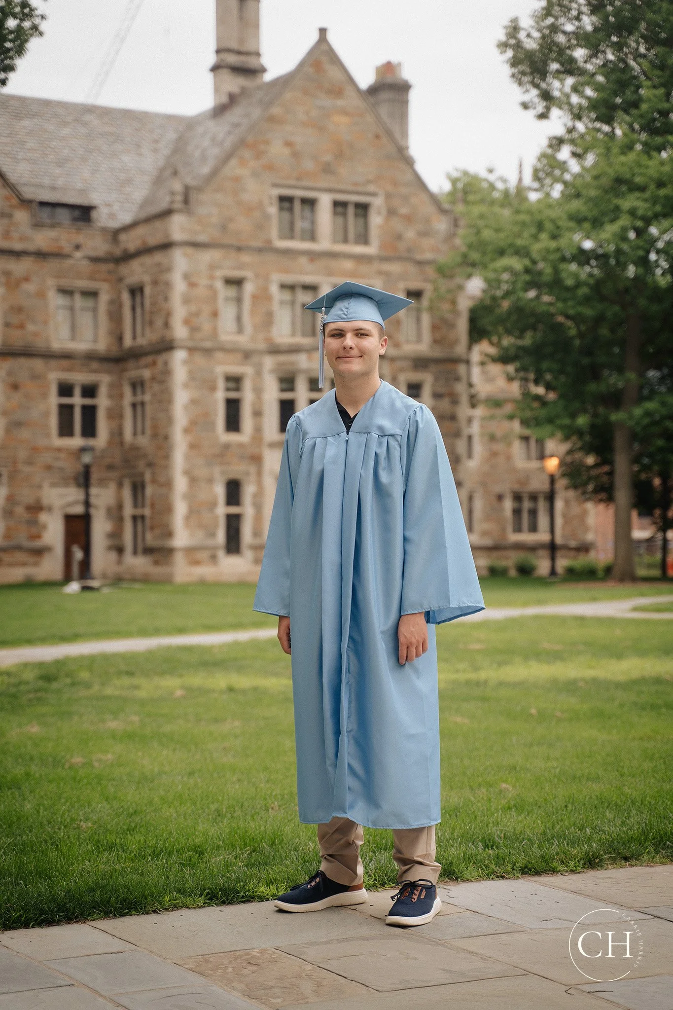 A young man in a light blue graduation gown and cap standing outdoors on a sidewalk in front of a large historic stone building with multiple windows and green trees.