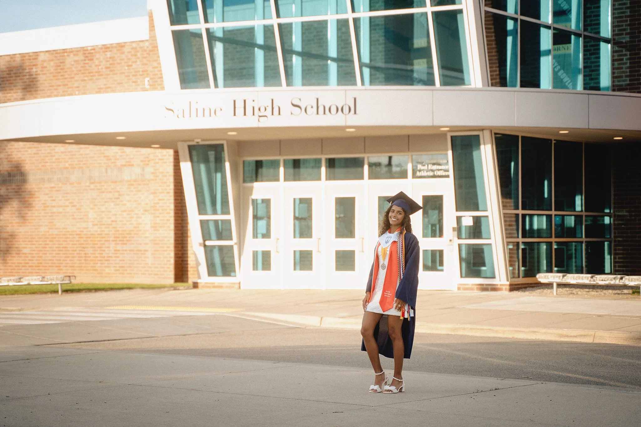 A young woman in graduation attire standing in front of Saline High School building.