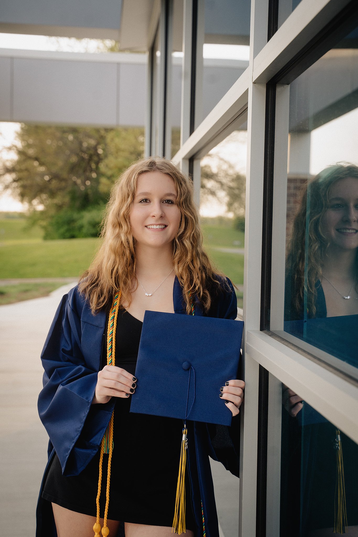 A young woman in a graduation gown holding a diploma and standing outside near a building with glass windows, smiling at the camera.