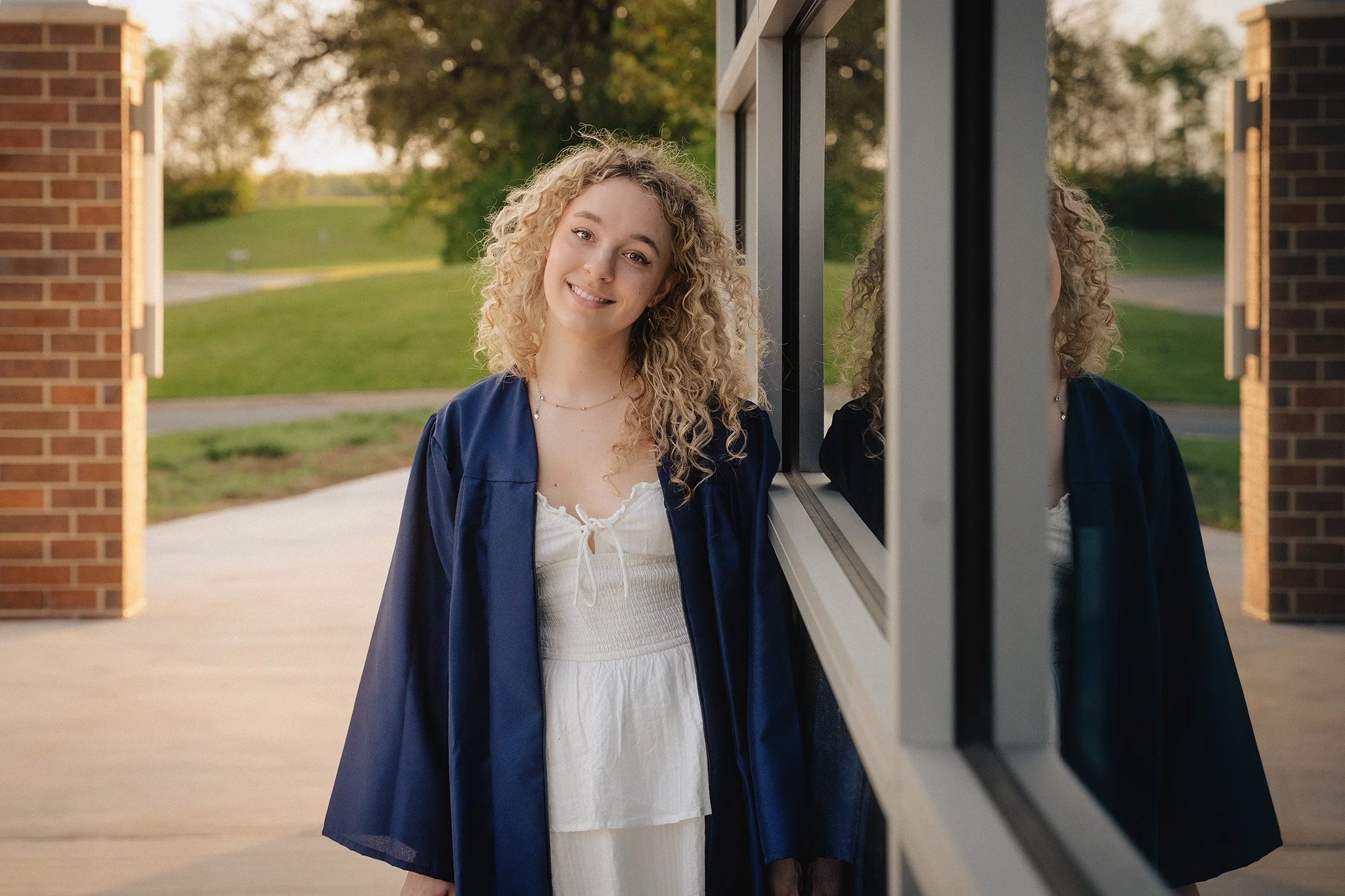 A young woman with curly blonde hair smiling at the camera outside a building with a brick wall and large windows.