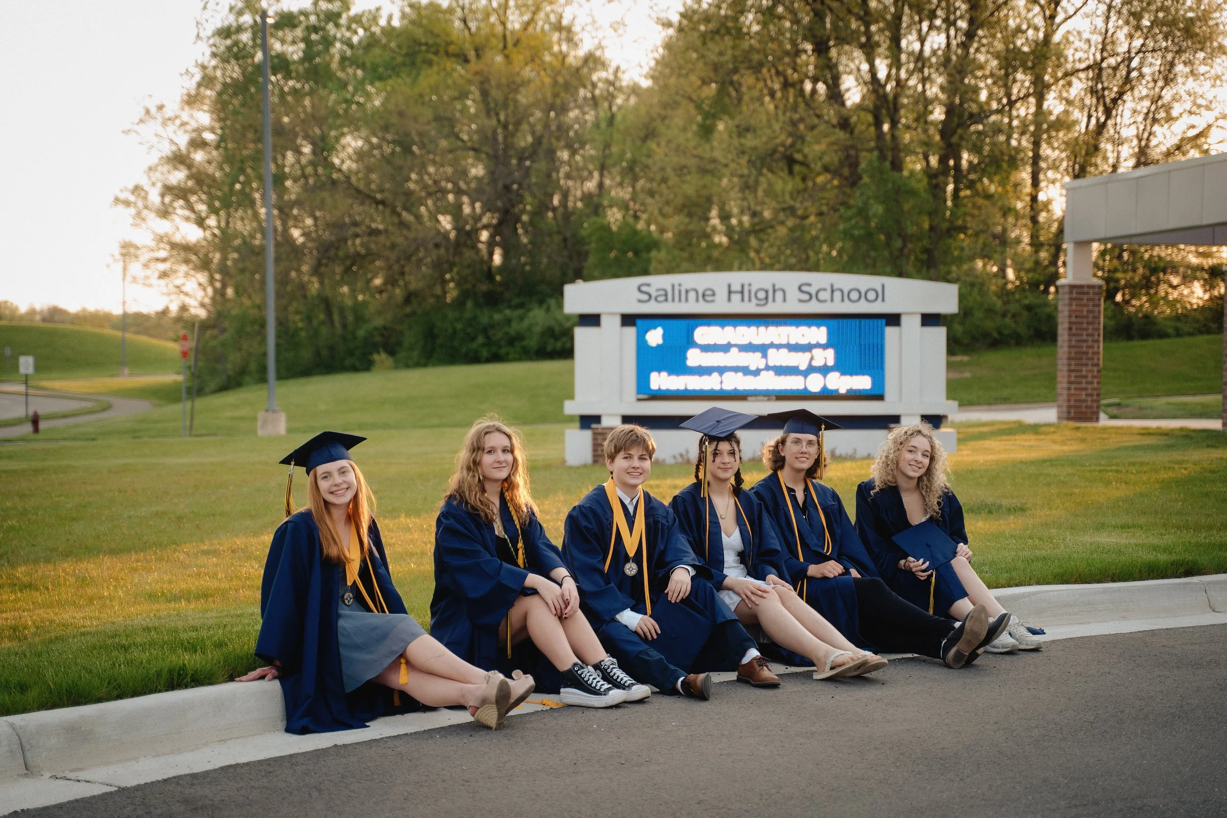 A group of six diverse high school graduates in blue caps and gowns sitting on the curb outside facing the camera, with a Saline High School sign and trees in the background during sunset.