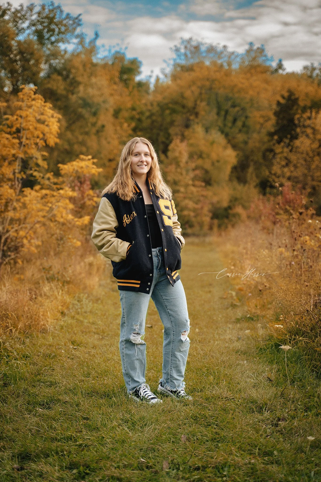 A young woman with wavy blonde hair smiling while standing outdoors on a grassy path surrounded by trees with autumn foliage. She is wearing a black and tan varsity jacket, ripped light blue jeans, and black and white sneakers.