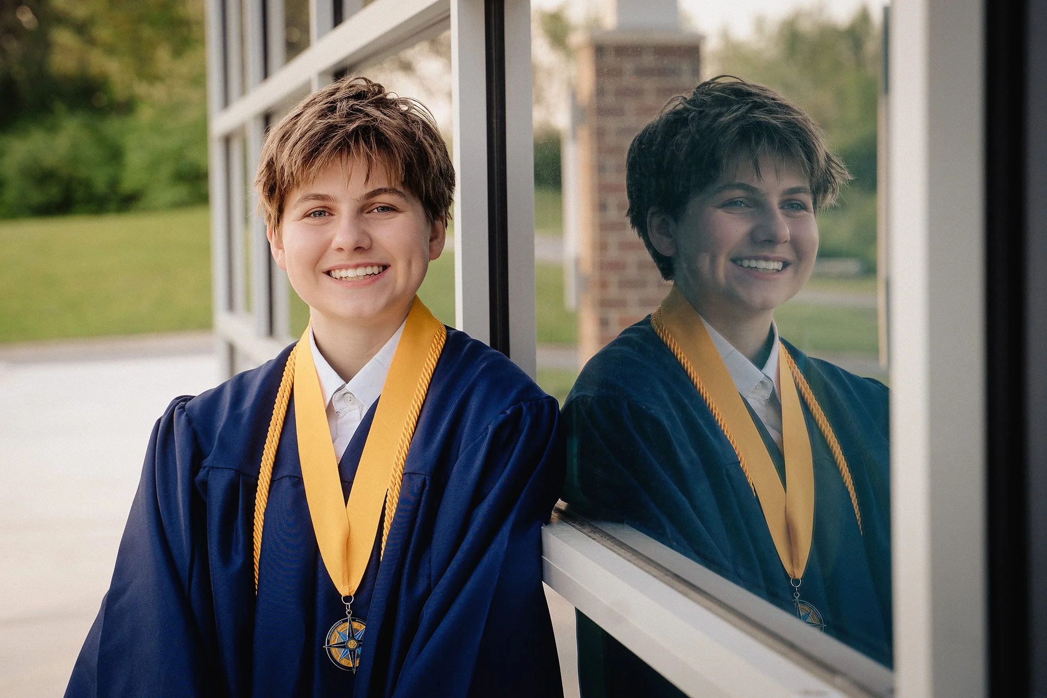 A young boy with brown hair, smiling in a graduation gown and medal, standing outside near a reflective window.