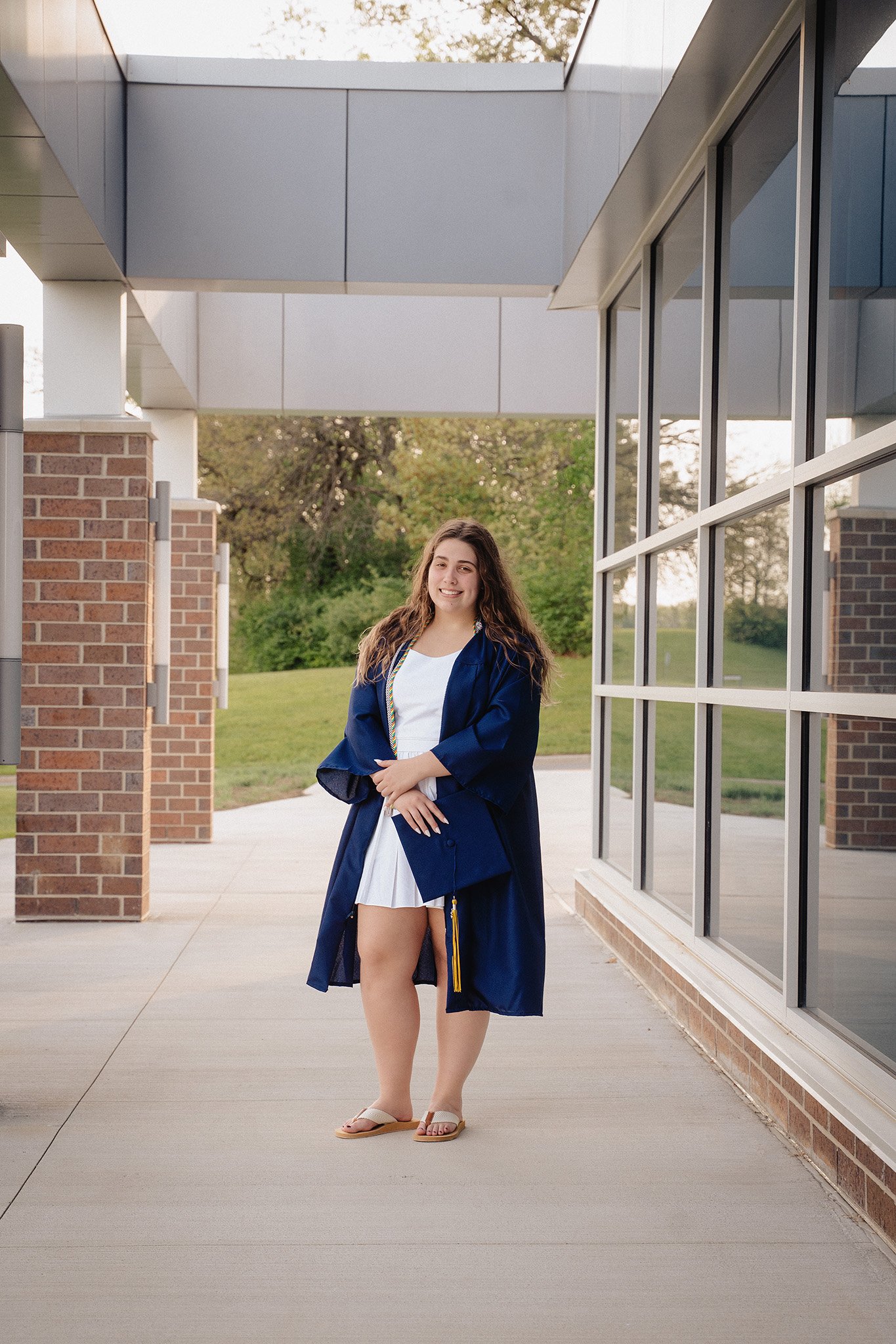 A young woman in a white dress and blue graduation gown stands outdoors in front of a modern building with large windows and a brick wall. She is smiling and wearing sandals.