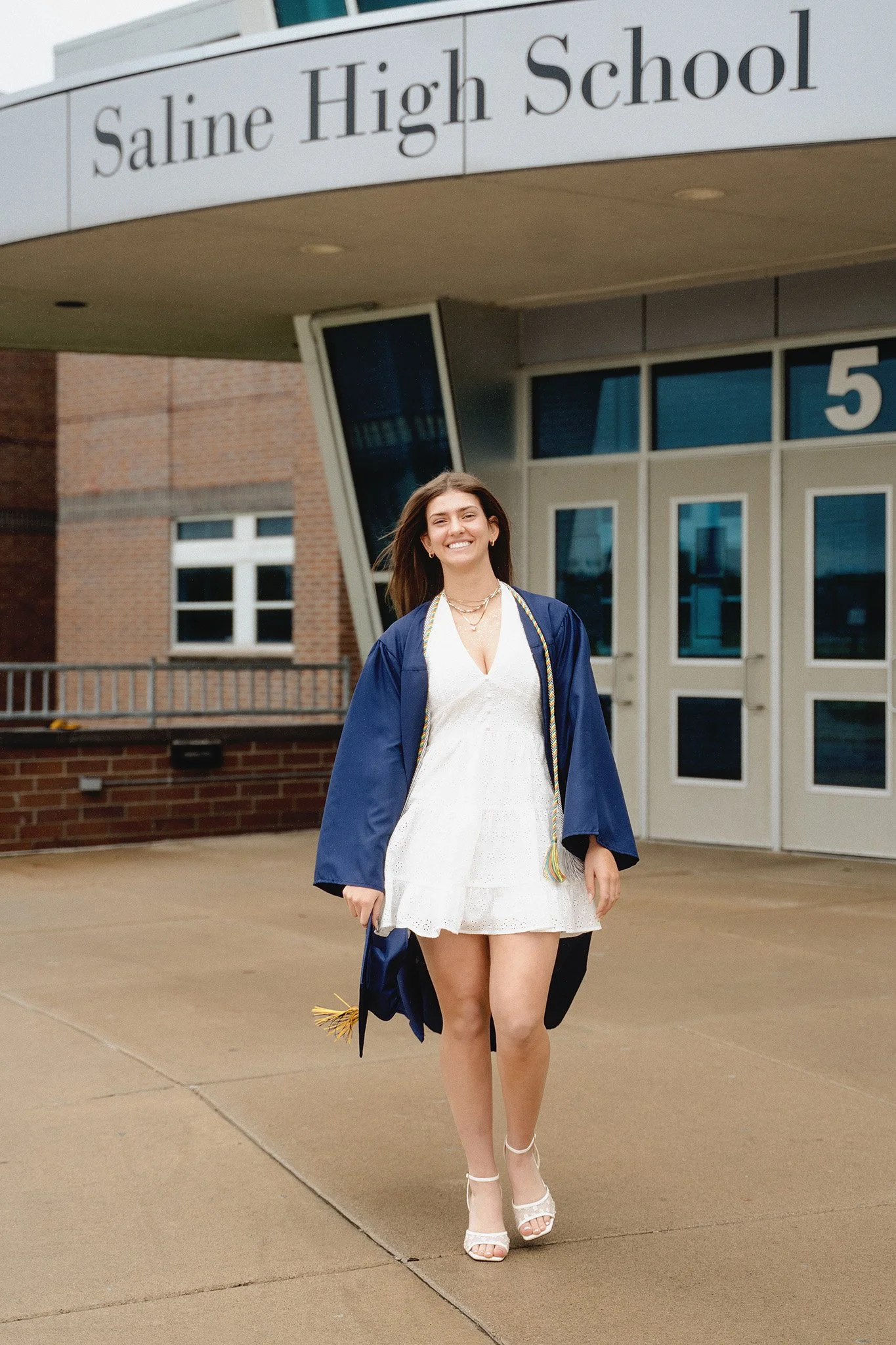 A young woman in a white dress and white high heels proudly wearing a blue graduation gown and cap with a multicolored tassel, standing outside the entrance to Saline High School, smiling after graduation.