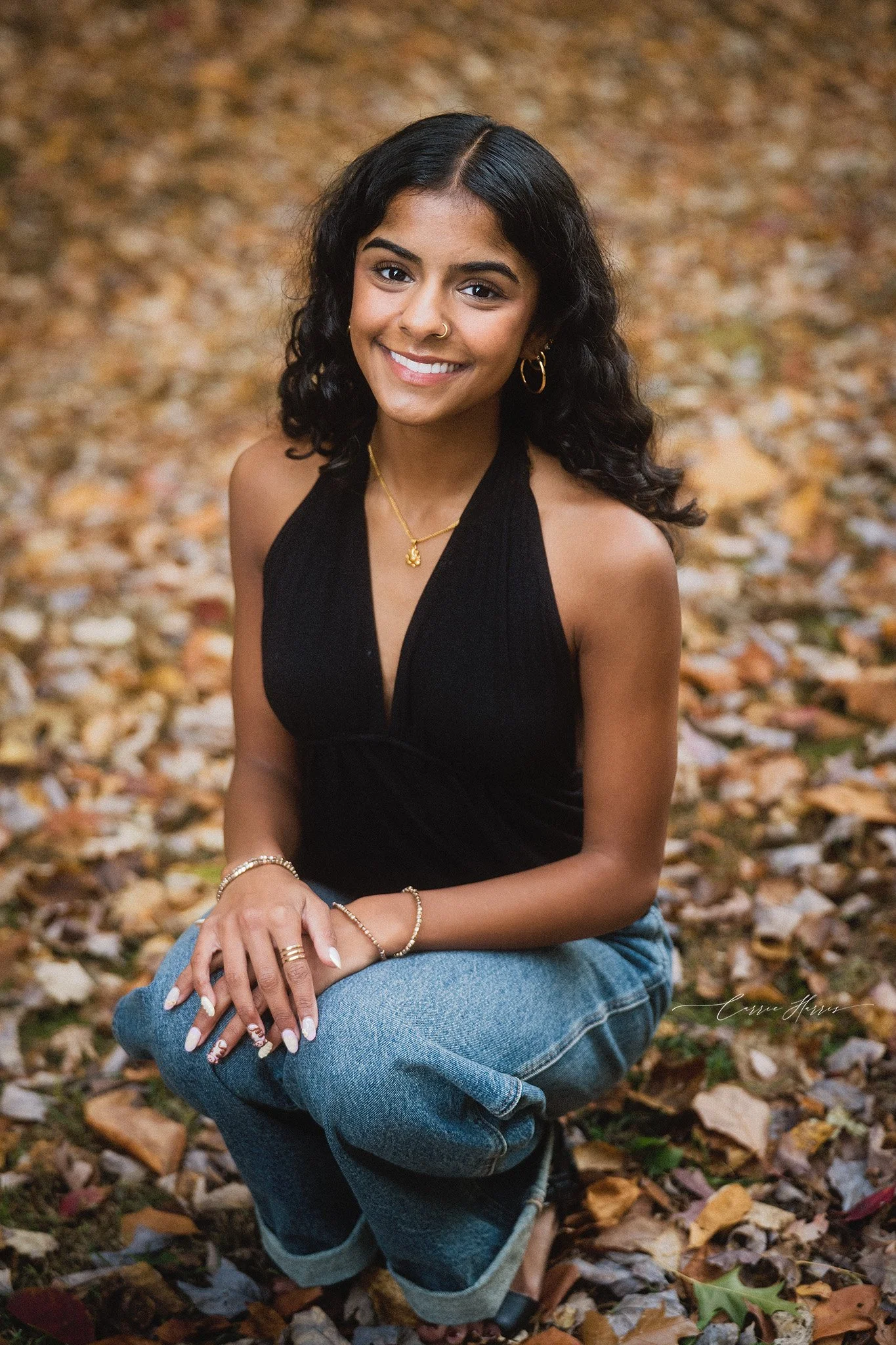 Young woman with curly black hair, wearing a black top and blue jeans, sitting on fallen autumn leaves.