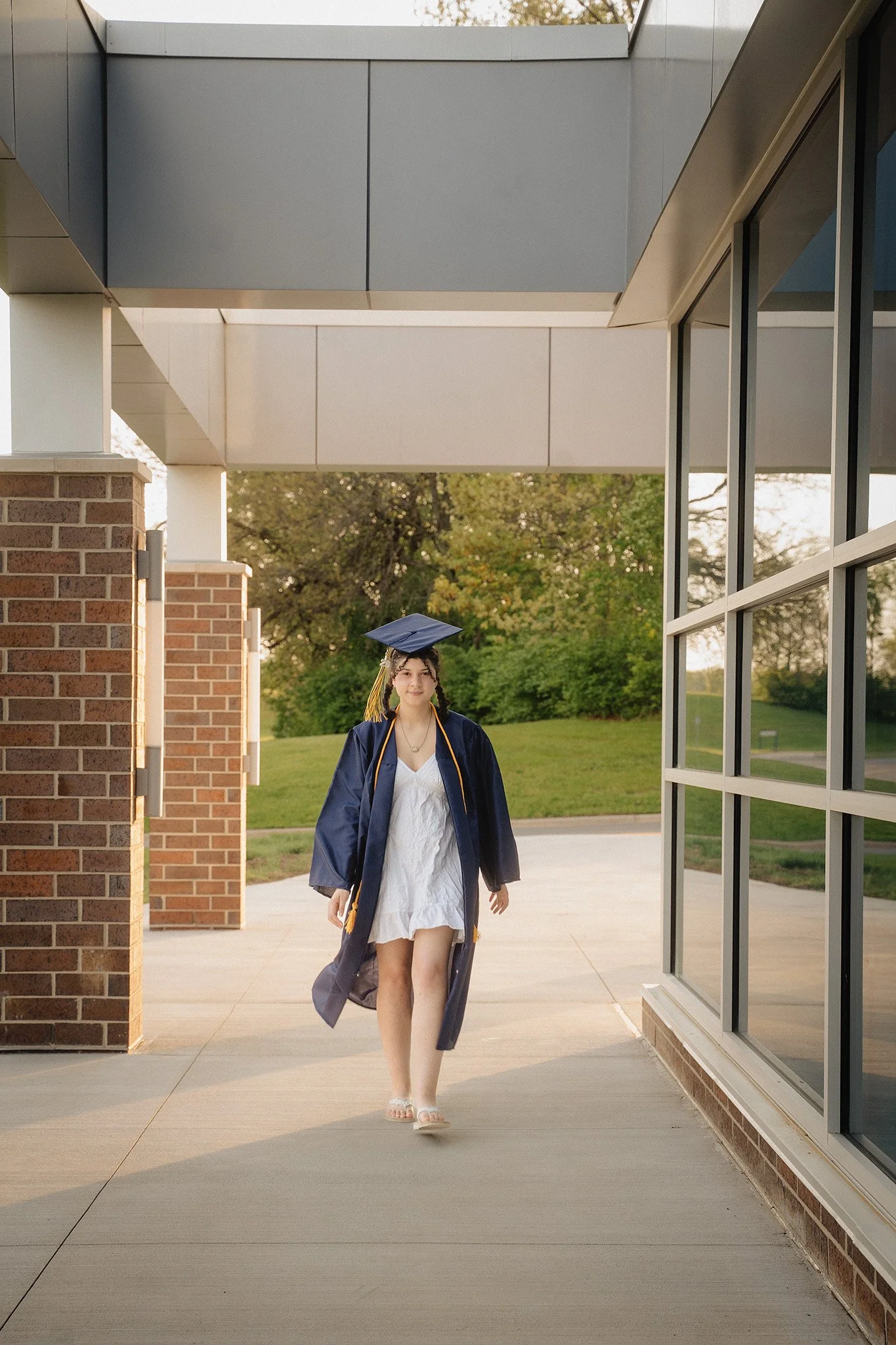 A young woman in a graduation gown and cap walking outdoors under a modern building structure with glass windows and brick pillars, green trees, and a grassy area in the background.