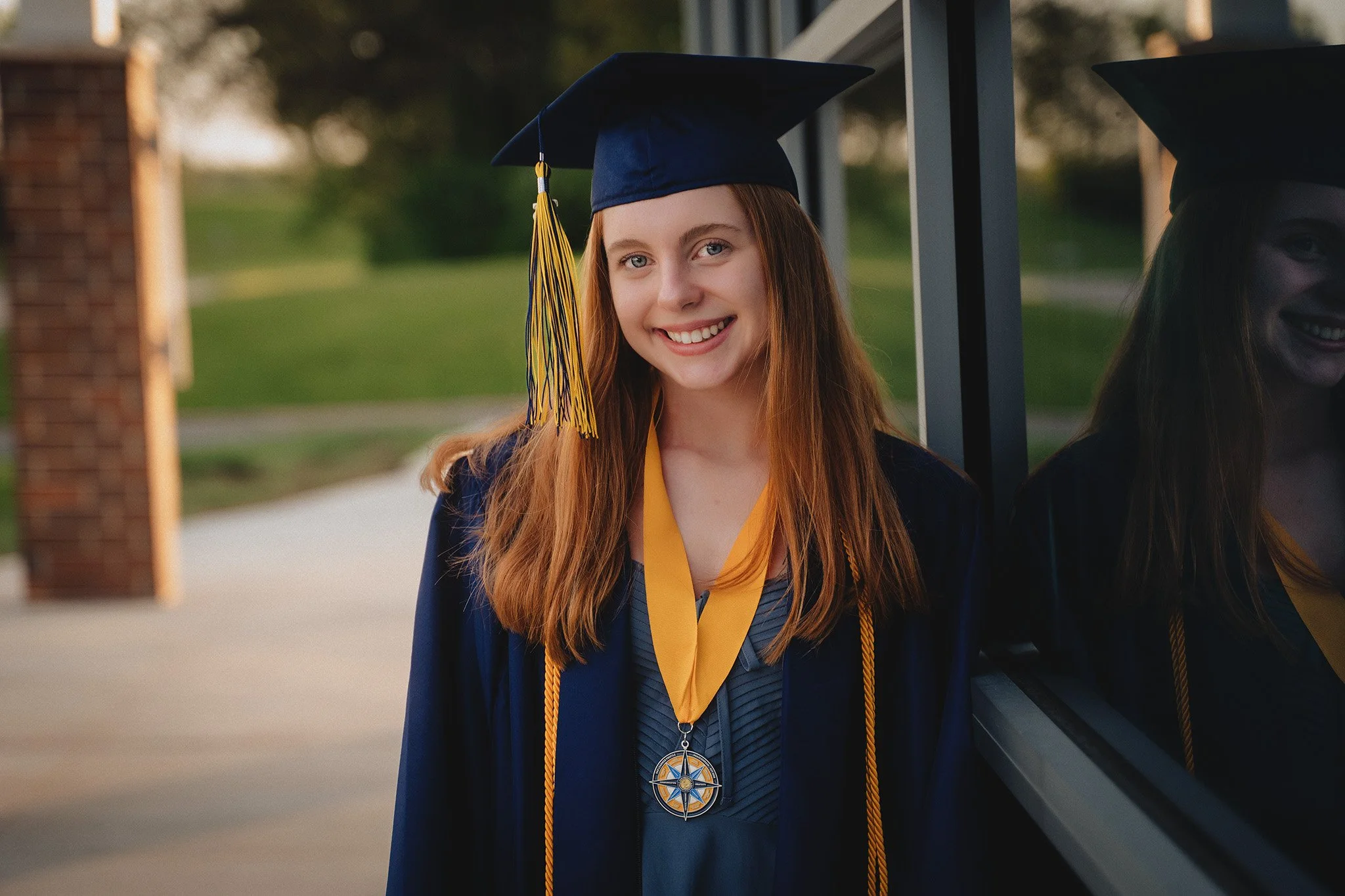 A young woman graduating outdoors, wearing a navy blue cap and gown, with a gold tassel and a medal hanging around her neck, smiling at the camera.