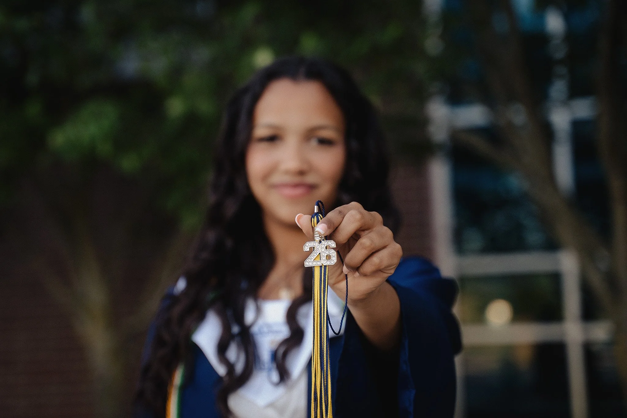 A young woman with dark, curly hair smiling and holding a graduation cap keychain with the number 26, wearing a blue graduation gown and multicolored cords, standing outdoors with blurred trees in the background.