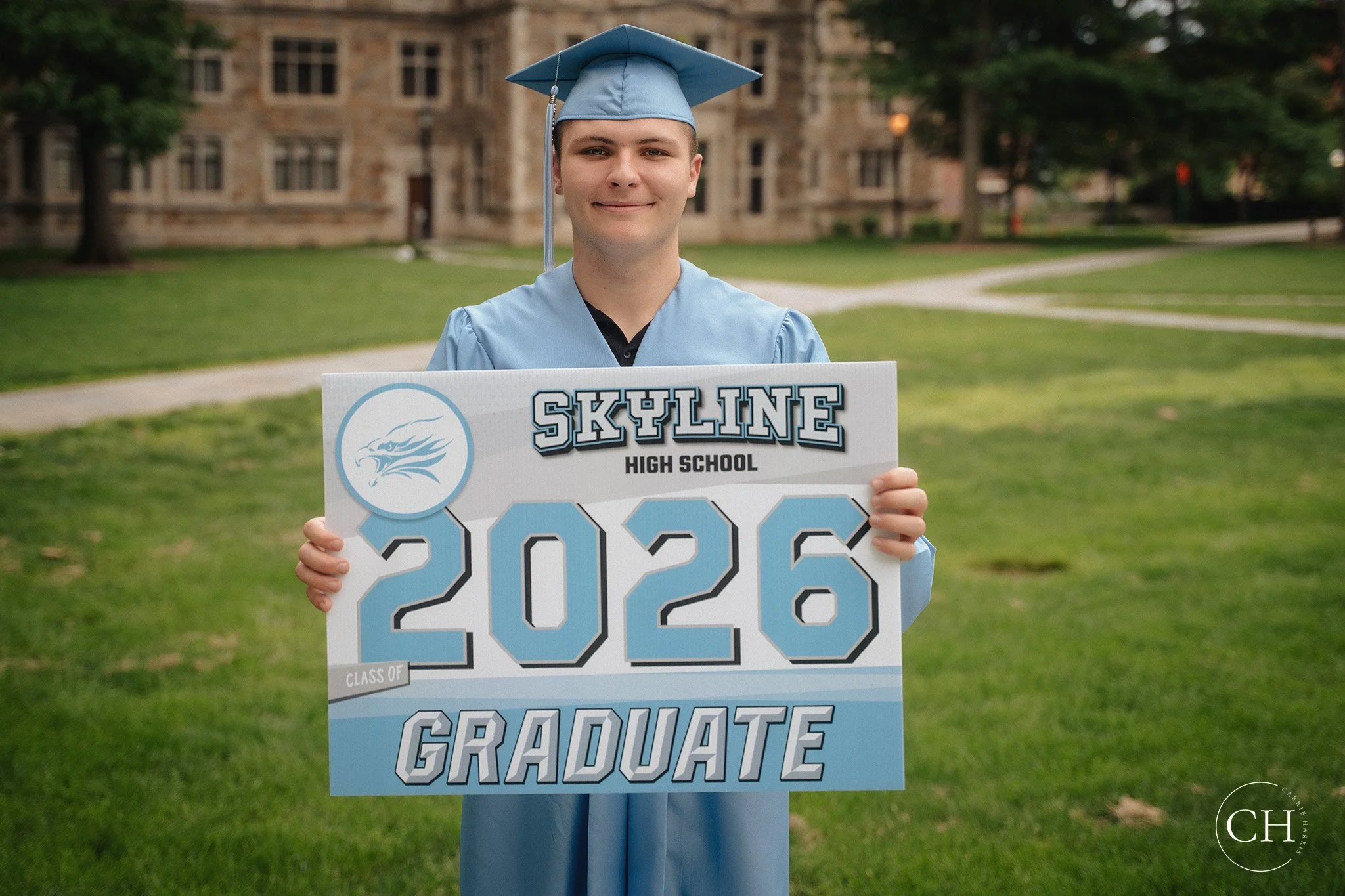 Graduating student in blue cap and gown holding a sign with 'Skyline High School' and 'Class of 2023' on it, standing on a grassy area with a historic building in the background.