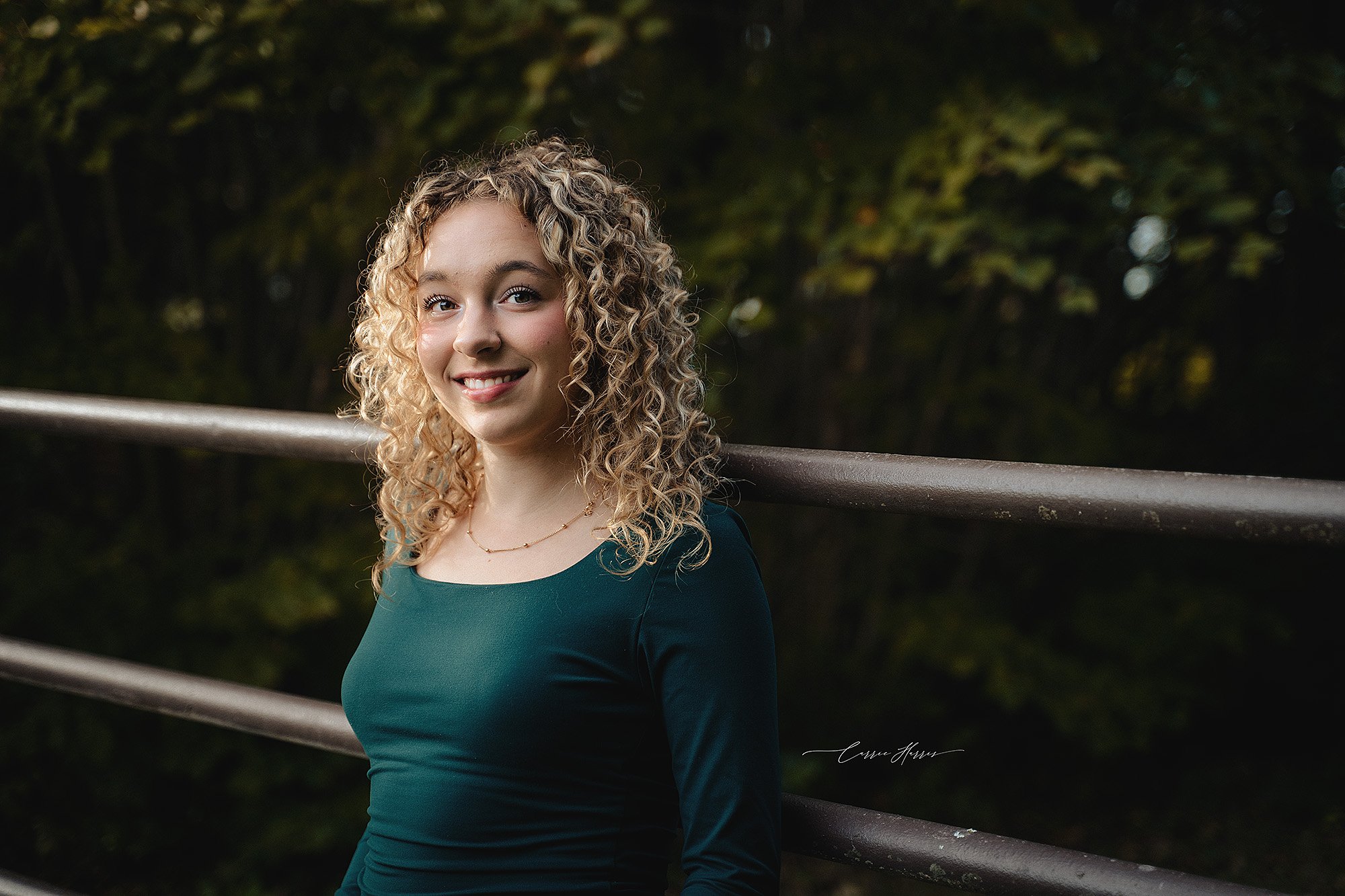A young woman with curly blonde hair smiling outdoors, wearing a dark green top and a delicate necklace, standing near metal railings with a blurred green leafy background.