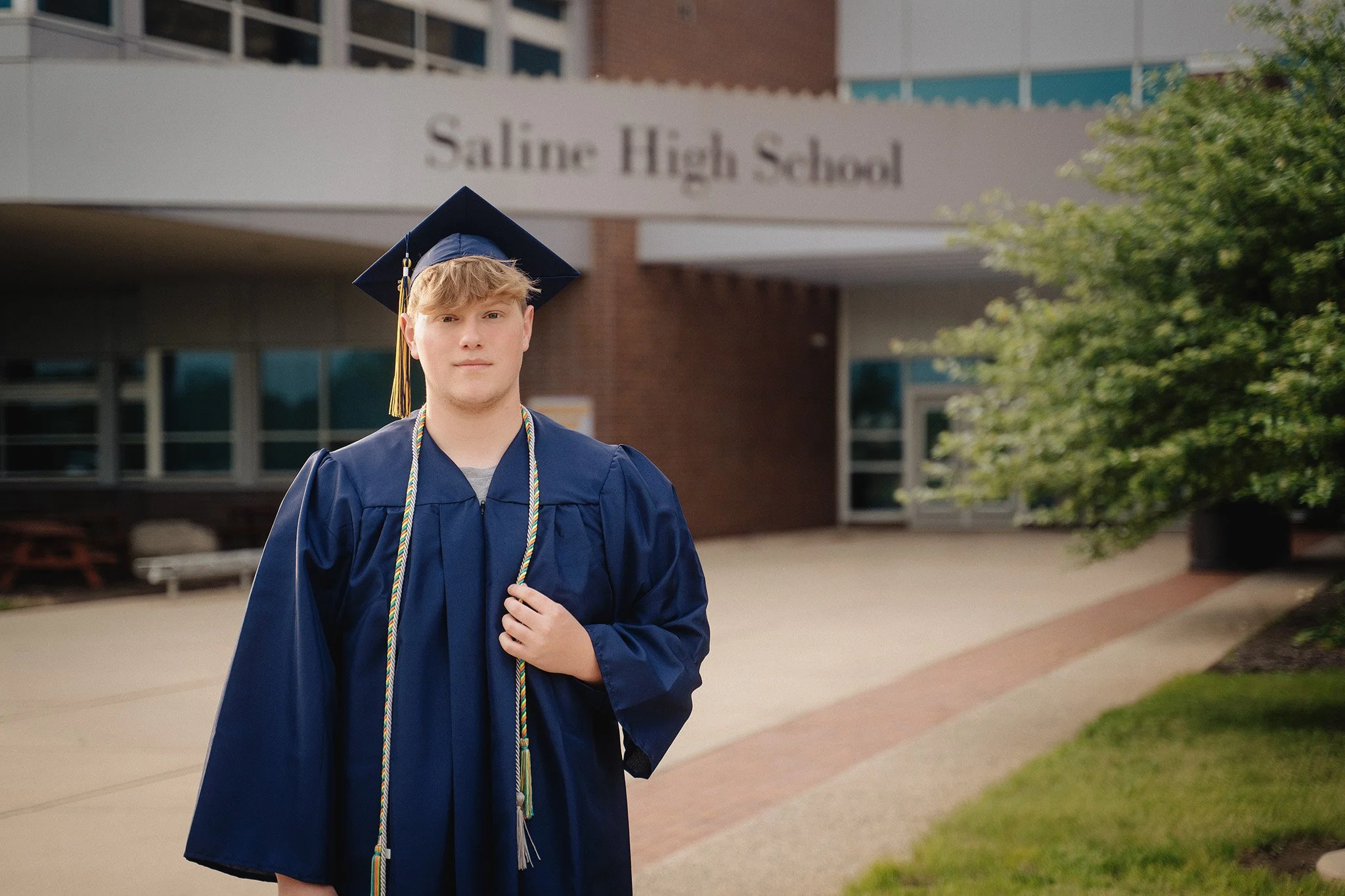 A young man in a blue graduation gown and cap standing outside in front of Saline High School with a blurred school building in the background.