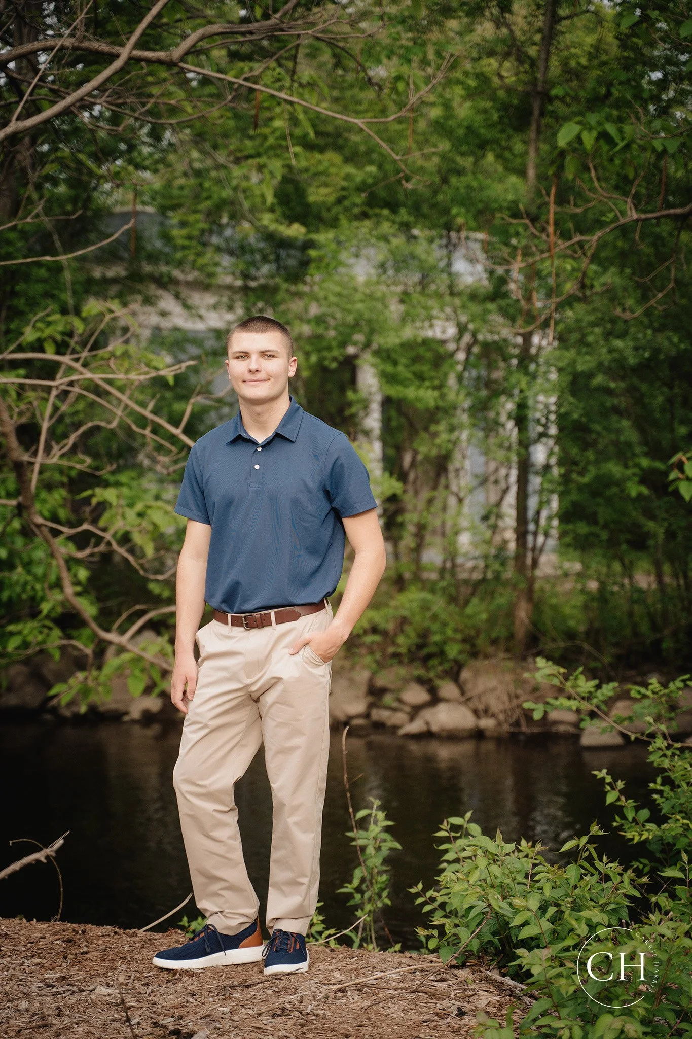 Young man standing outdoors near river, wearing blue shirt and tan pants, with trees and greenery in the background.