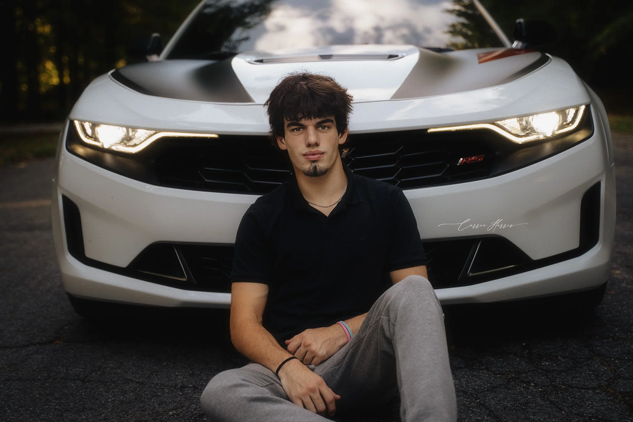 A young man with dark brown hair, a goatee, and a black shirt is sitting in front of a white sports car outdoors during the evening. The person is positioned directly in front of the car's front grille, with the car's headlights on and trees visible 