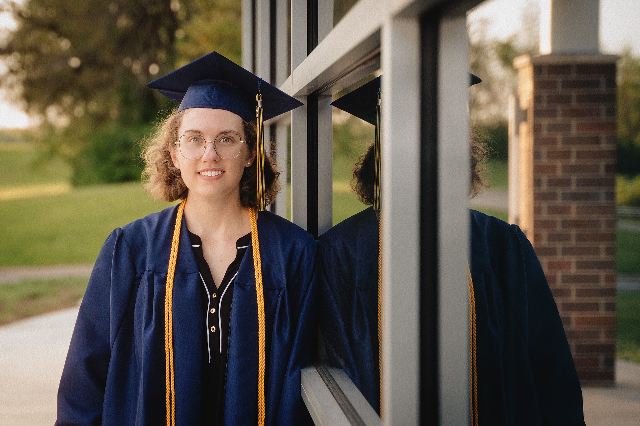 Graduating woman in cap and gown standing outside near a building, looking at the camera with a smile.