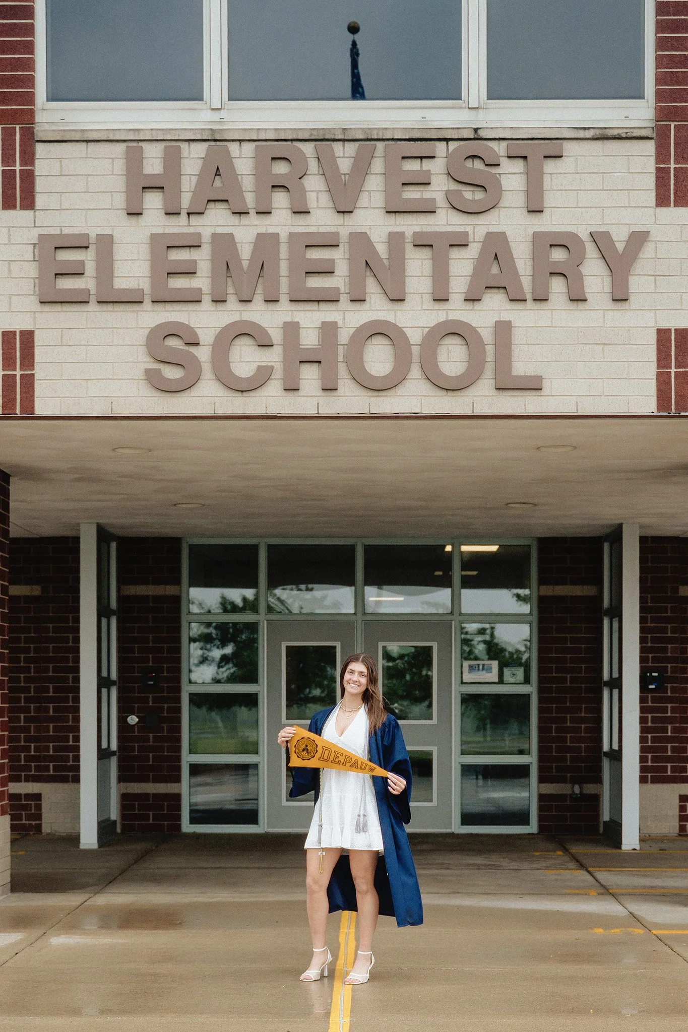 A young woman in a graduation cap and gown standing outside a school building, holding a diploma and smiling.