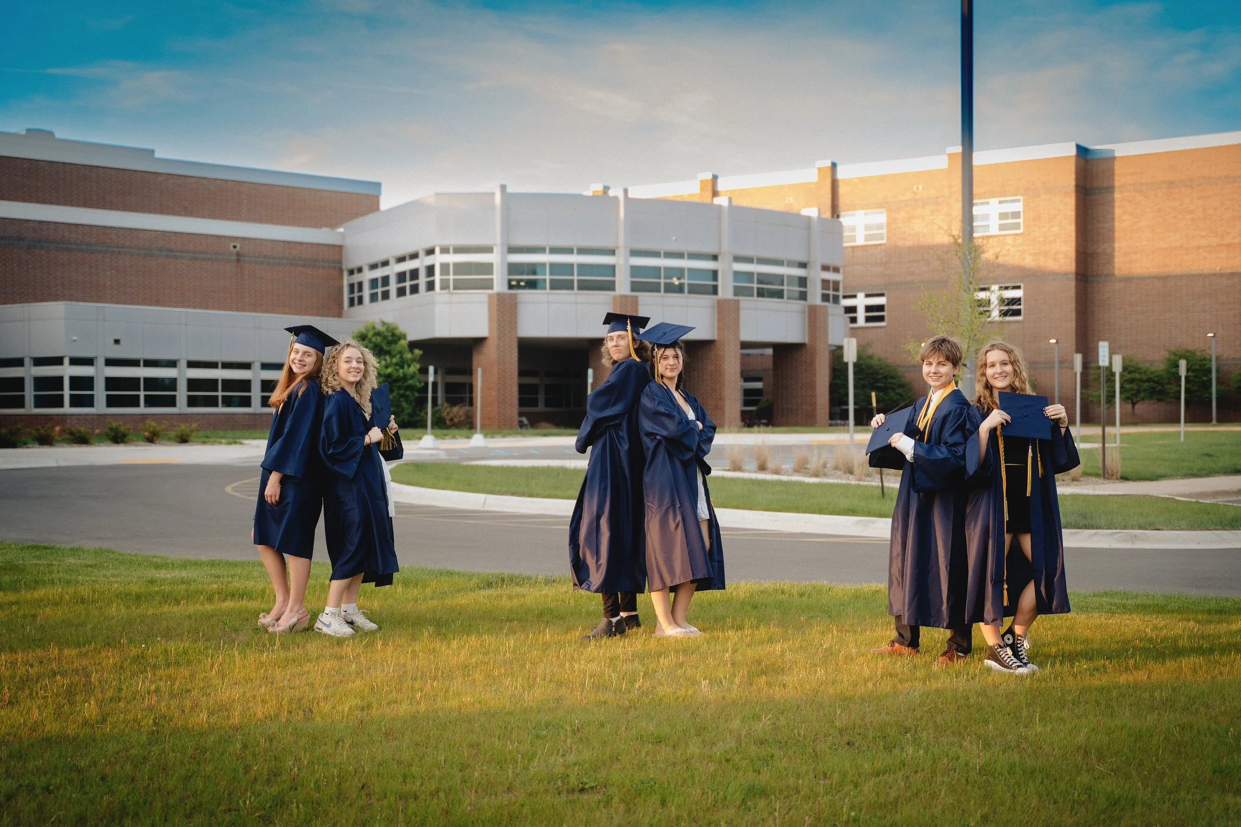 Group of five happy graduates in blue caps and gowns standing outside in front of a school building, holding their diplomas.