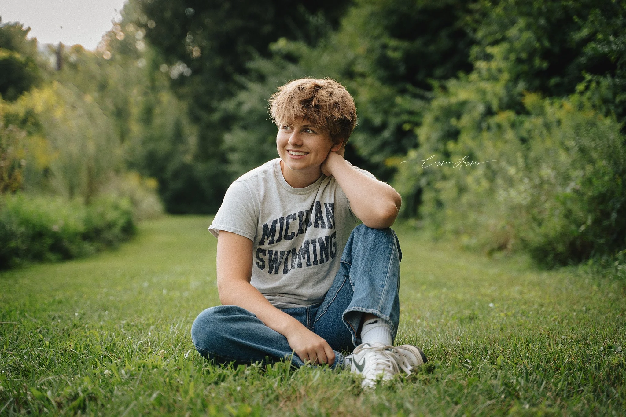 A young person with short hair sitting on the grass in a park, smiling, wearing a gray t-shirt with text, jeans, and white sneakers, surrounded by green trees.