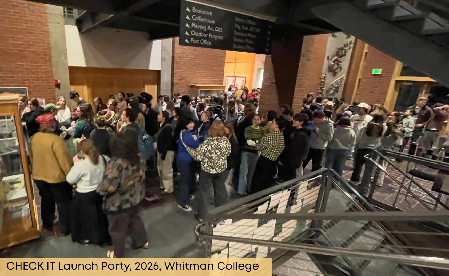 A large crowd of people gathered inside a building with brick walls during the CHECK IT Launch Party at Whitman College in 2026. The scene shows a busy event with individuals chatting near the entrance, some standing in line, and others mingling. Signs hanging from the ceiling direct attendees to various locations such as the bookstore, coffeehouse, meeting room, outdoor program, and post office.
