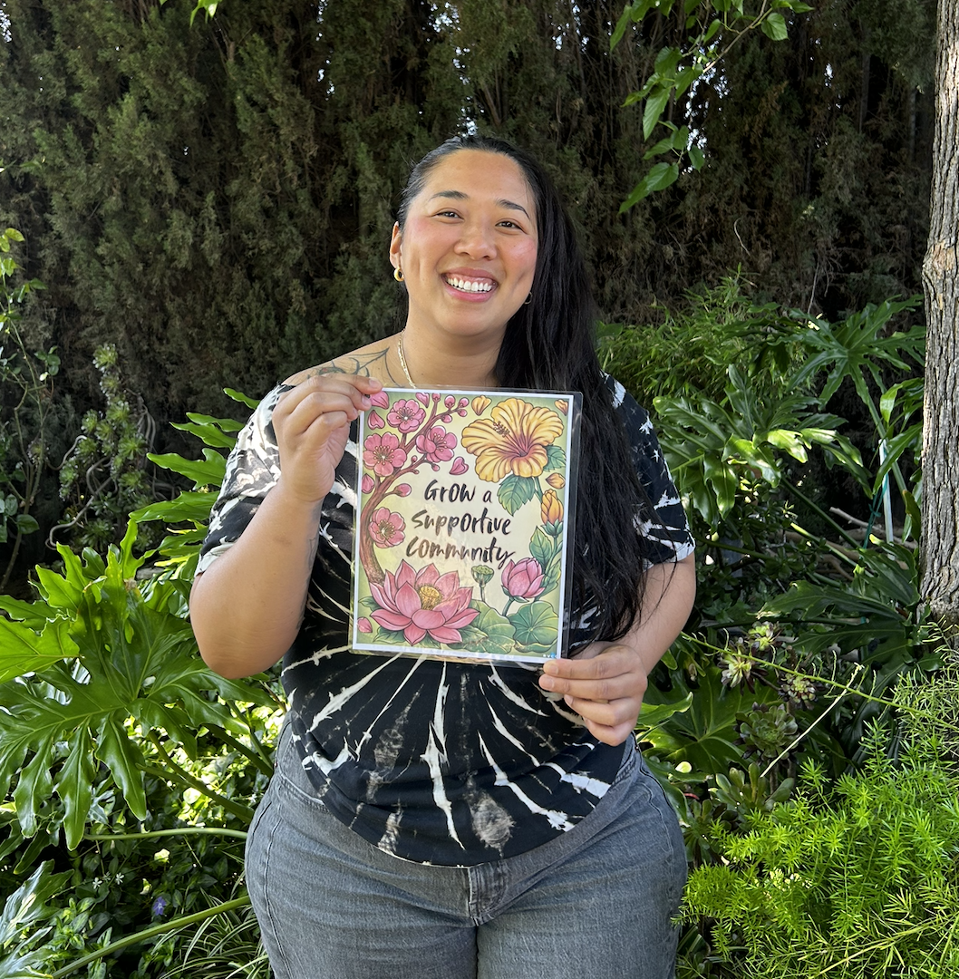 Woman holding a colorful sign that says 'Grow a Supportive Community' in a lush garden with green foliage.
