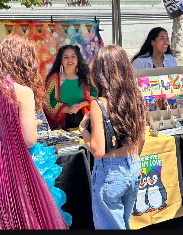 Group of women talking at a booth with colorful rainbow decorations and artwork, outdoors in sunlight.