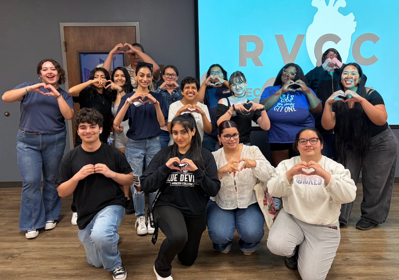 Group posing after a sexual violence bystander intervention workshop, making heart shapes with their hands, in front of a large screen displaying a logo with a heart and the letters R V C C.
