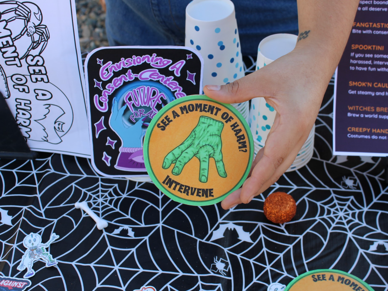 Person holding a coaster with a green hand and the text "See a moment of harm? Intervene". The background includes Halloween-themed decorations, like spiderwebs and small skeleton stickers, on a black tablecloth with white web patterns.
