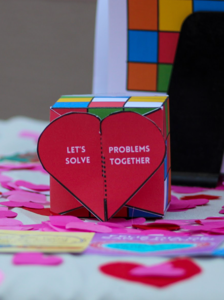 A heart-shaped paper craft with the message 'Let's Solve Problems Together' written on it, surrounded by pink paper hearts and other colorful craft materials for TDVAM and Valentine's Day.