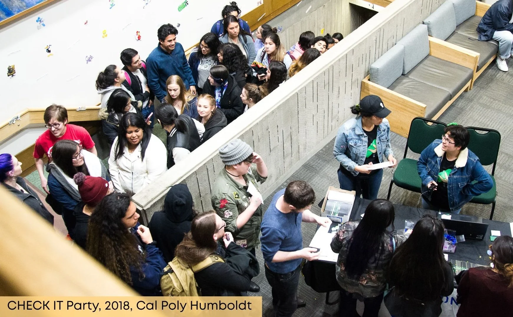 A large group of young adults gather in a hallway, some standing in lines and others waiting at a table to participate in activities, at the CHECK IT Party 2018 at Cal Poly Humboldt.
