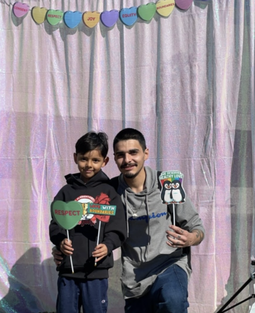 A father and son are posing together at a healthy relationships pop-up fair photobooth. They are holding photobooth props that say consent and "we deserve healthy relationships"