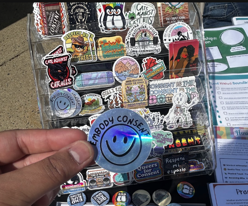 Collection of violence prevention themed stickers, with someone holding up the holographic "embody consent" smiley sticker.