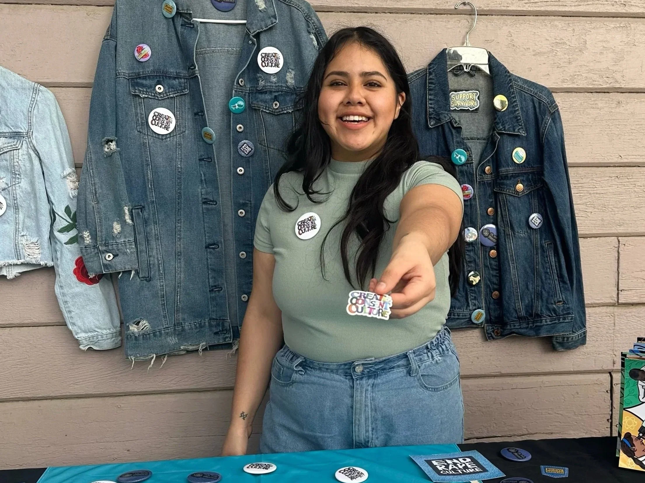 A young woman with long dark hair smiling in front of a display wall covered with denim jackets decorated with various pins and patches. She is wearing a light green t-shirt and high-waisted jeans. The wall behind her is made of horizontal wooden panels.
