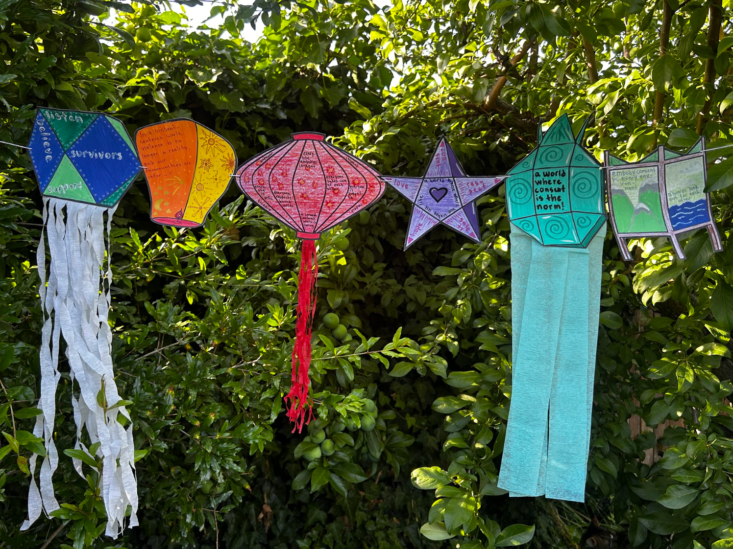 Colorful paper lanterns and stars hanging on a string in front of green foliage, decorated with words and drawings related to peace, love, and support.