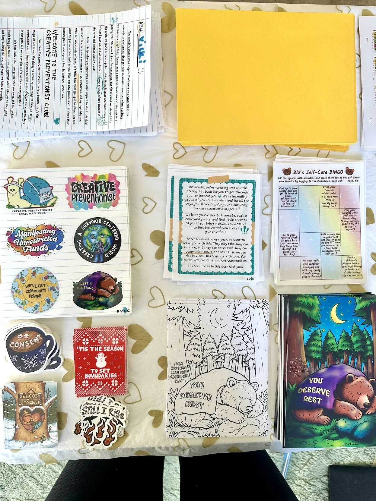 A table covered with various colorful printed materials, including stickers, coloring pages, cards, and flyers, some with messages about rest and community, on a beige tablecloth with gold hearts.