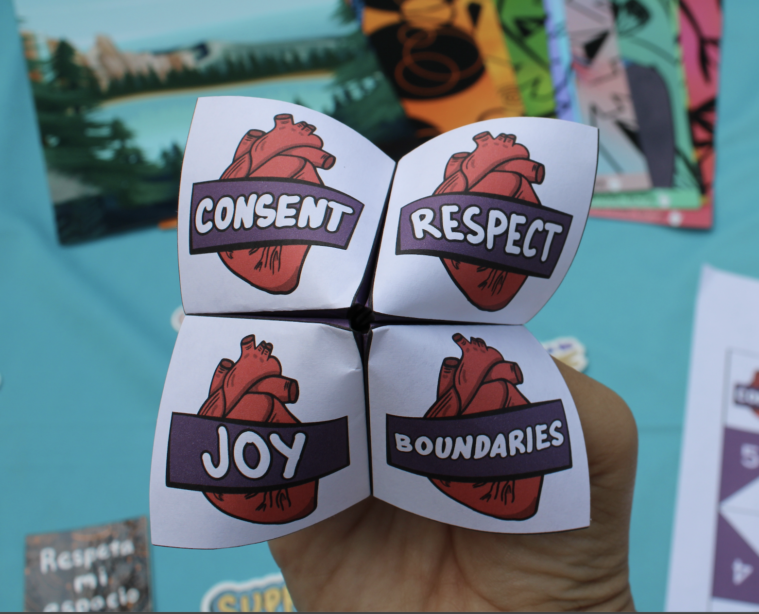 A DVAM themed origami chatterbox, fortune teller, cootie catcher featuring a drawing of a human heart with purple banners on each square reading "Consent," "Respect," "Joy," and "Boundaries."