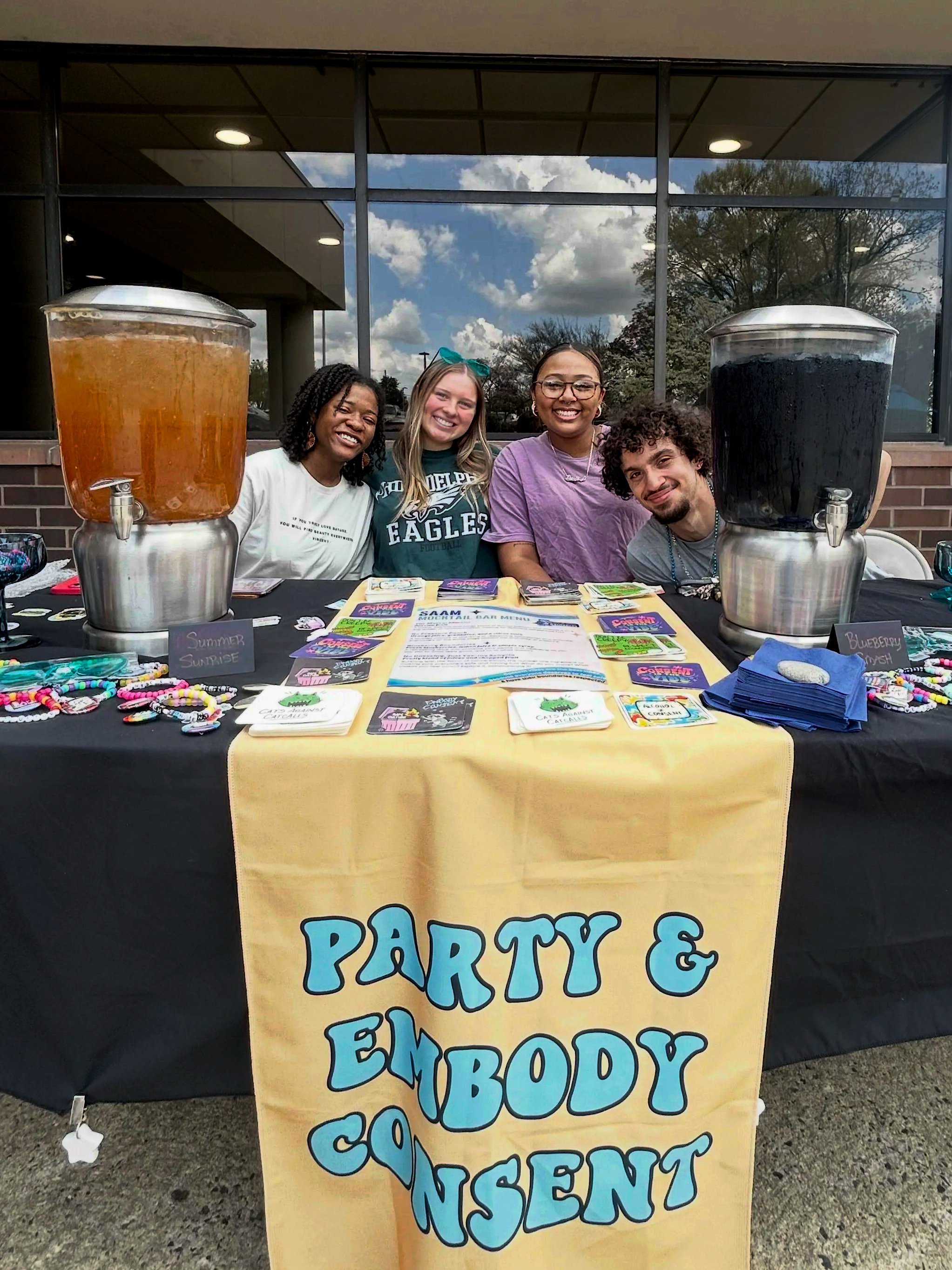A group of four smiling people sitting behind a consent-centered mocktail bar featuring sexual violence prevention bracelets, coasters, and menu.