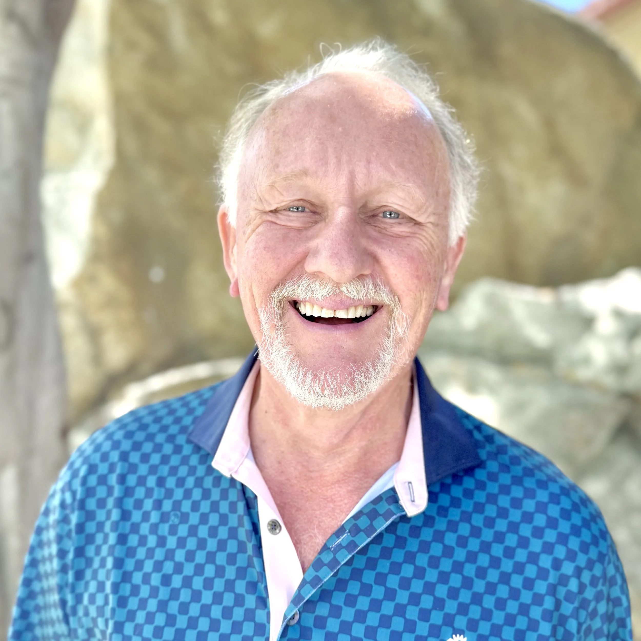 Smiling elderly man with white hair and beard, wearing a blue patterned shirt, standing outdoors with a blurred stone background.