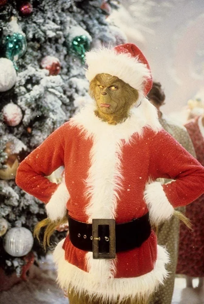 A photo of Jim Carey's Grinch wearing a Santa suit, standing in front of a Christmas tree with his hands on his hips frowning
