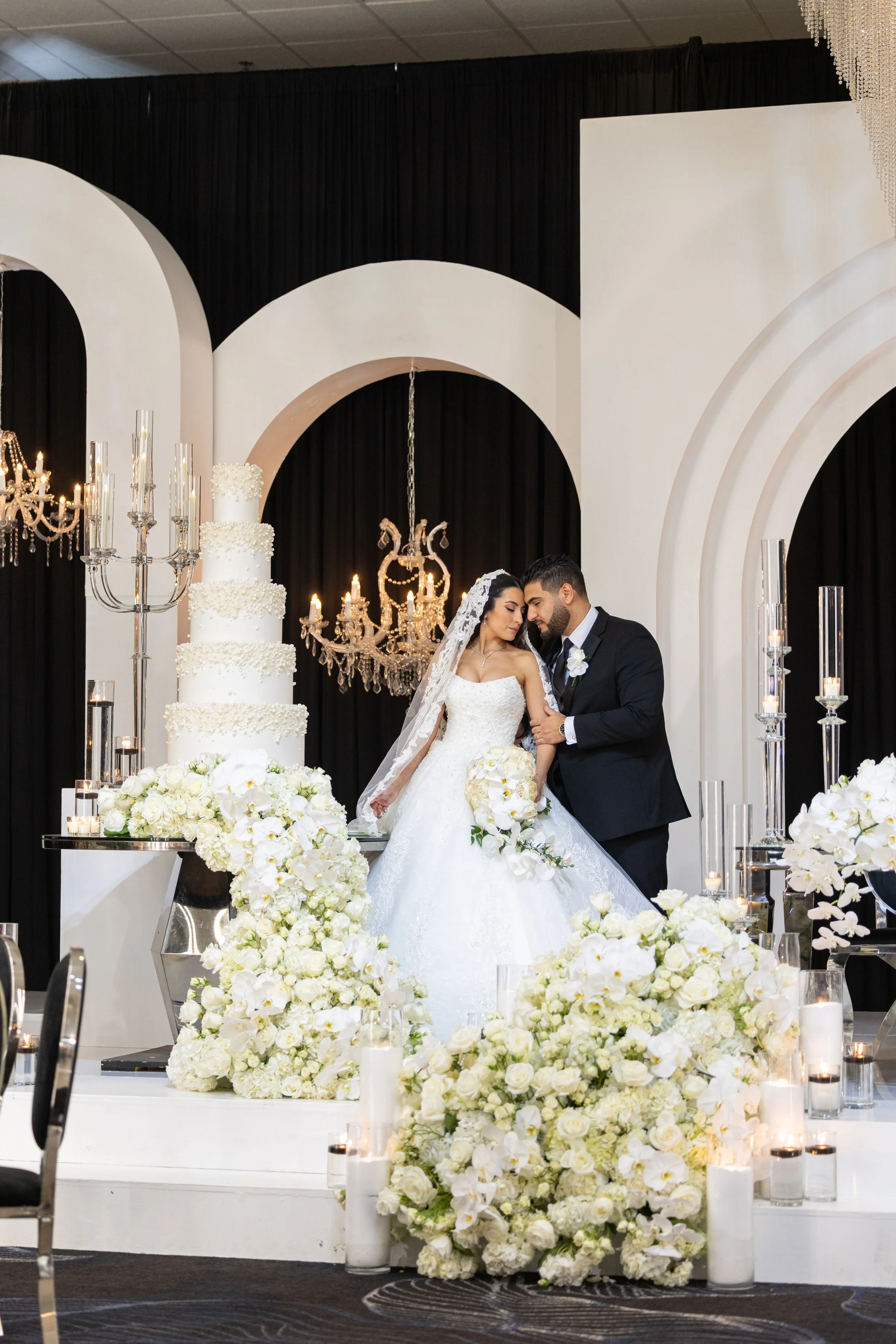 Bride and groom beside a tiered white wedding cake under arches and chandeliers