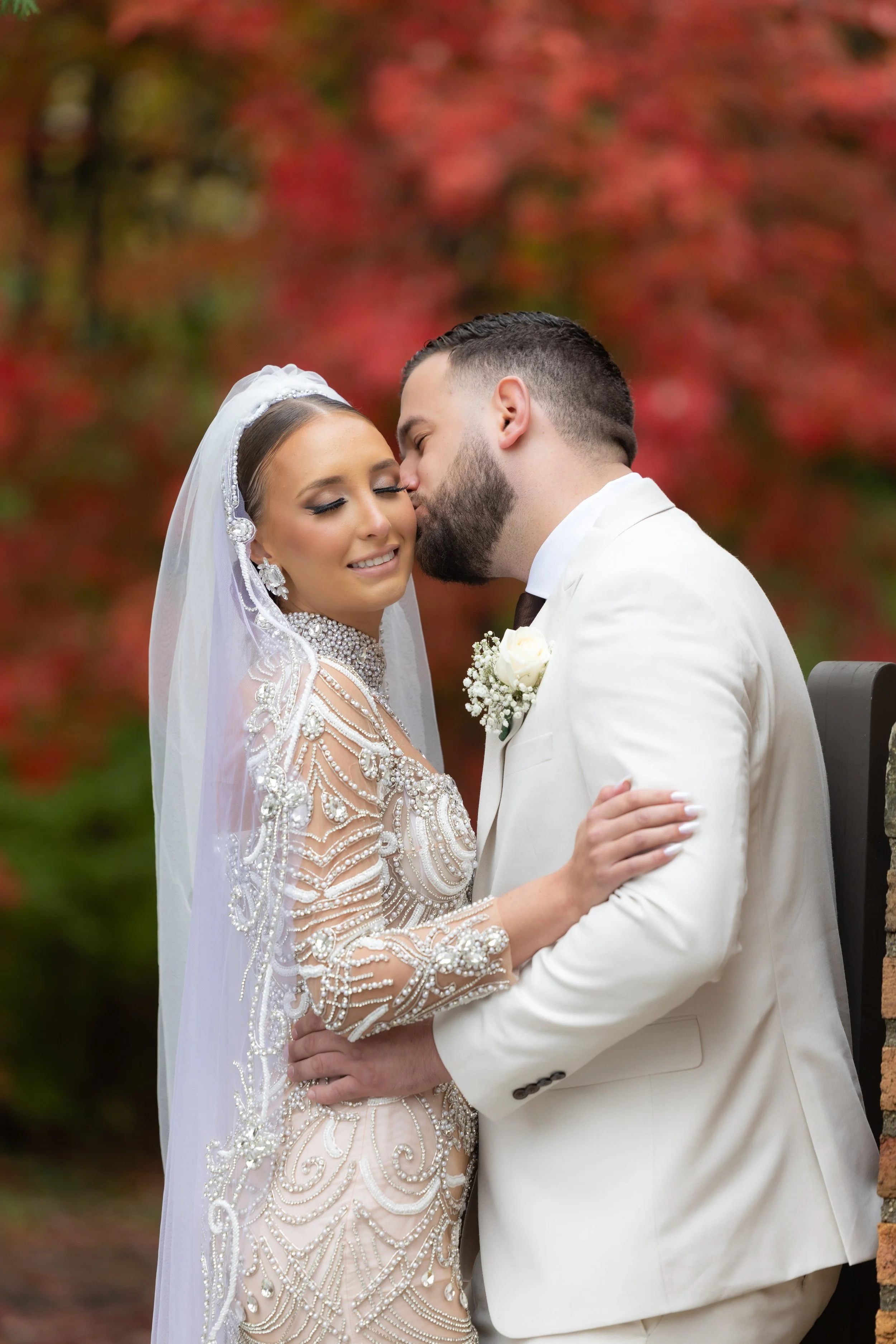 Groom kissing his bride's cheek in front of red autumn leaves