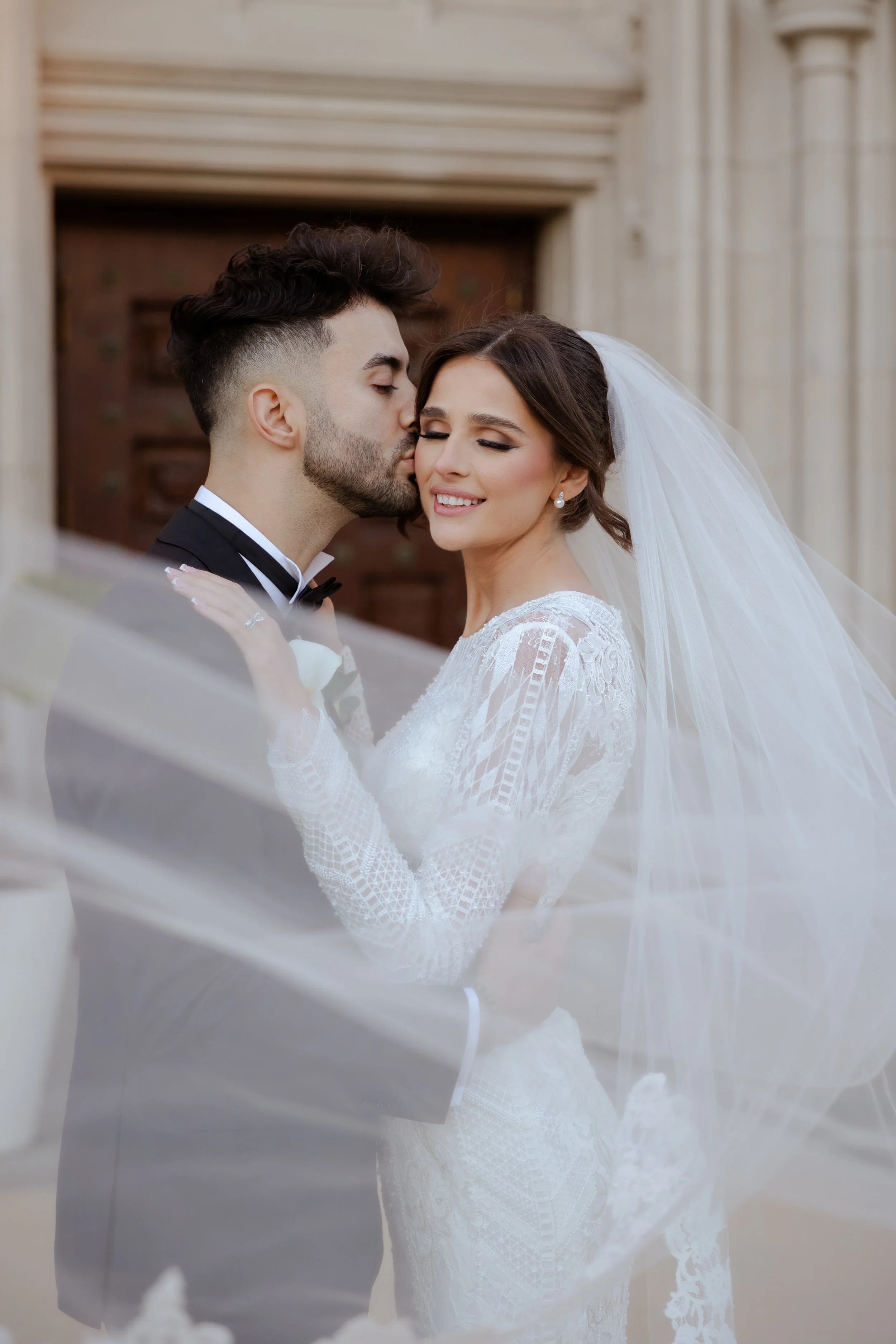 Groom kissing his bride's cheek as her veil drifts in the breeze