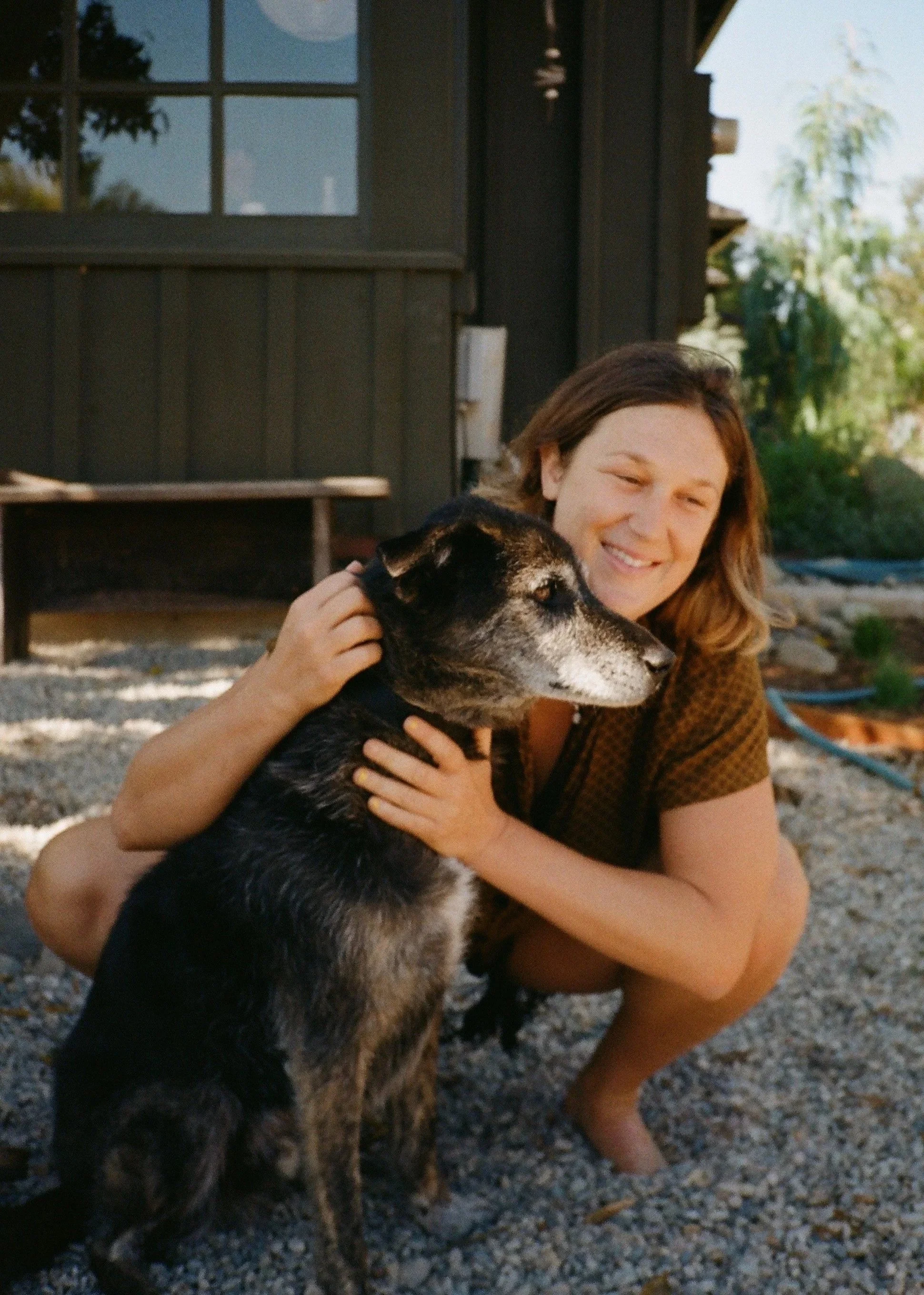 A woman hugging a black and tan dog outdoors on a gravel surface near a black wooden building with a window. The woman is smiling, and there are trees and other outdoor items in the background.