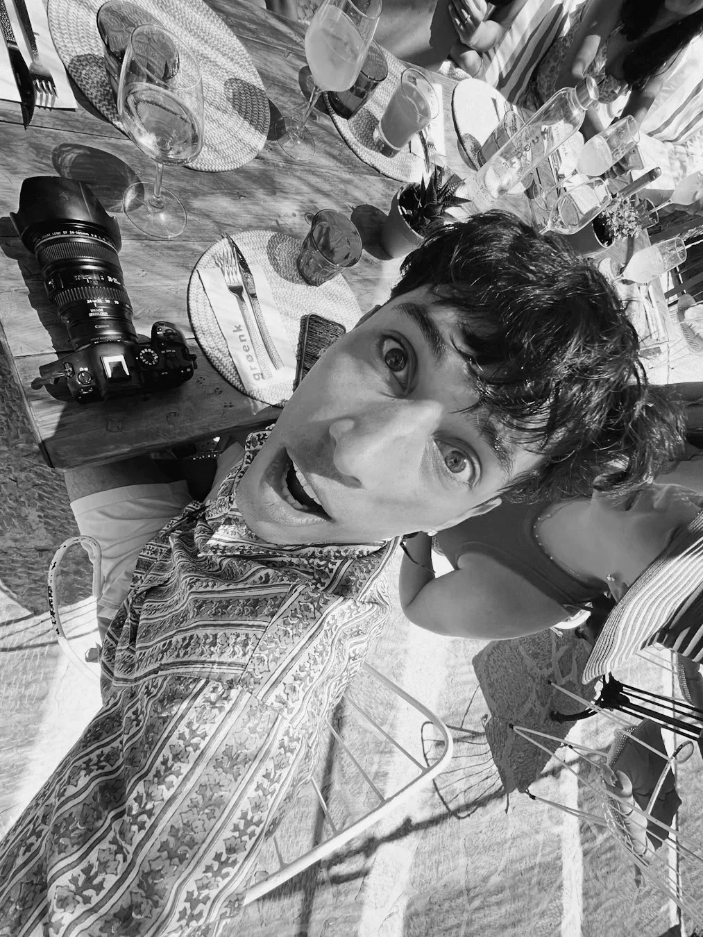 A young man with curly hair and a patterned shirt taking a selfie at an outdoor table set with plates, glasses, and drinks. There is a camera on the table and other people in the background.