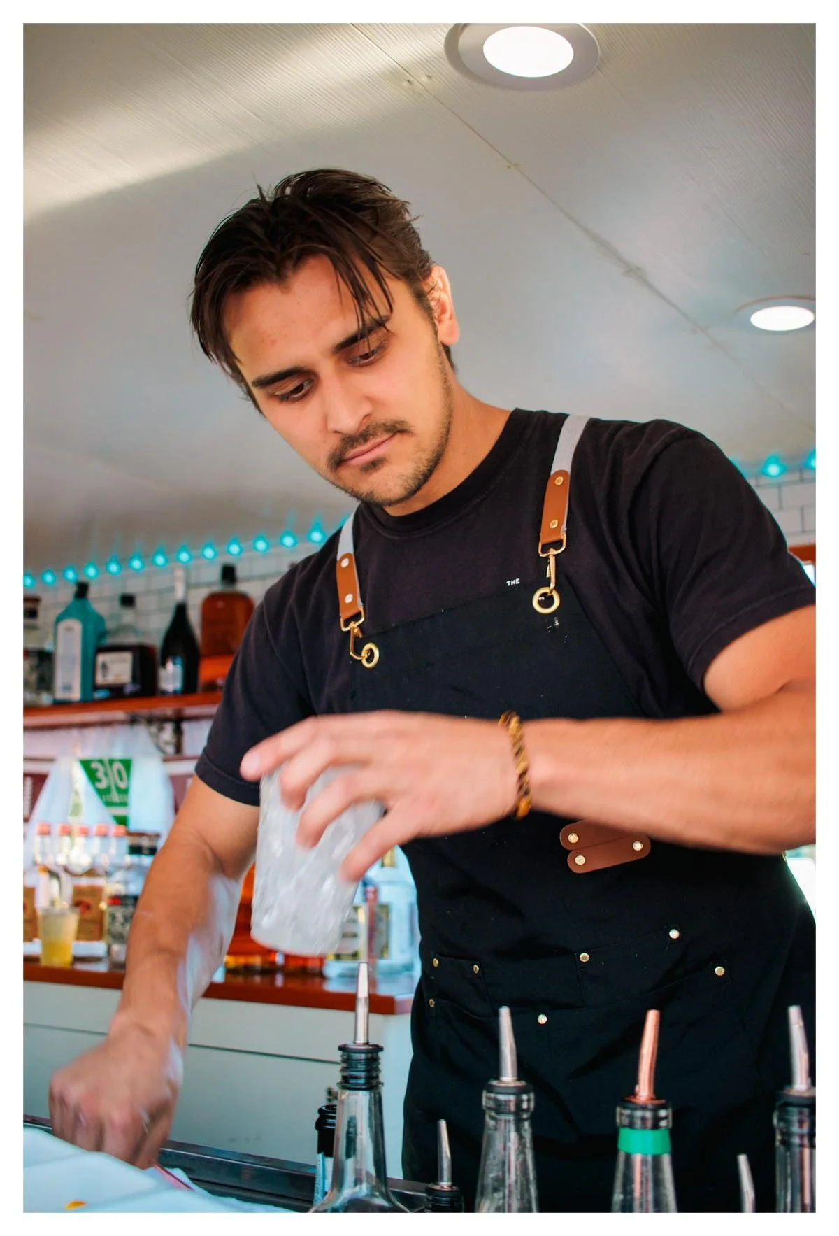 A young man with dark hair and a beard, wearing a black t-shirt and an apron, pouring a clear liquid from a container into a squeeze bottle in a casual restaurant or cafe setting.