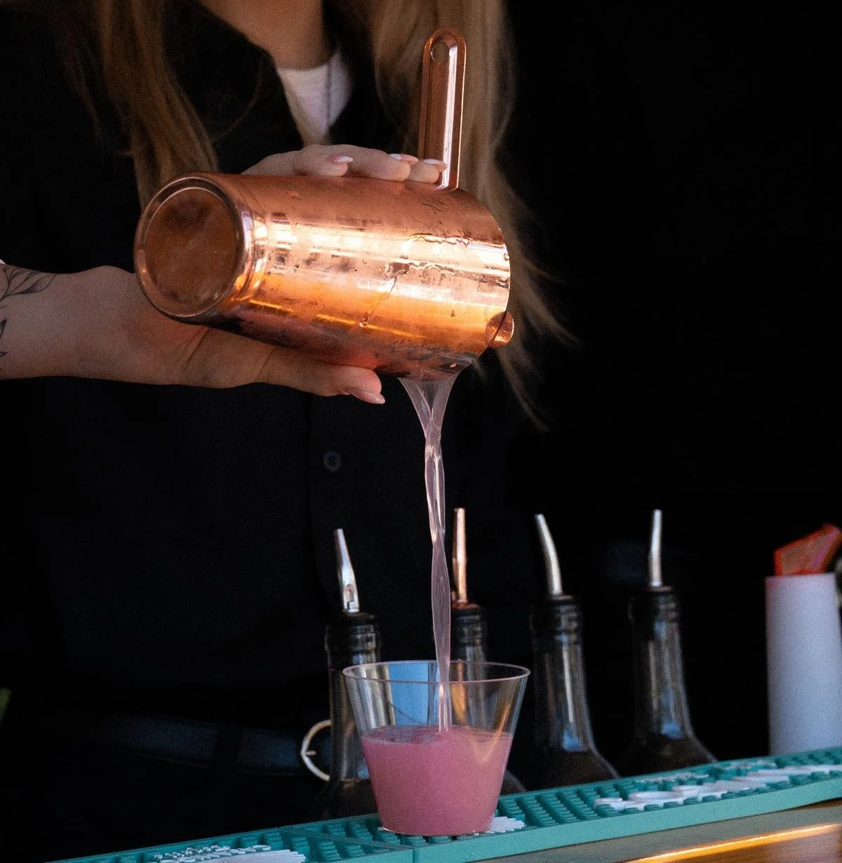 A bartender pours pink liquid from a copper cocktail shaker into a glass at a bar.