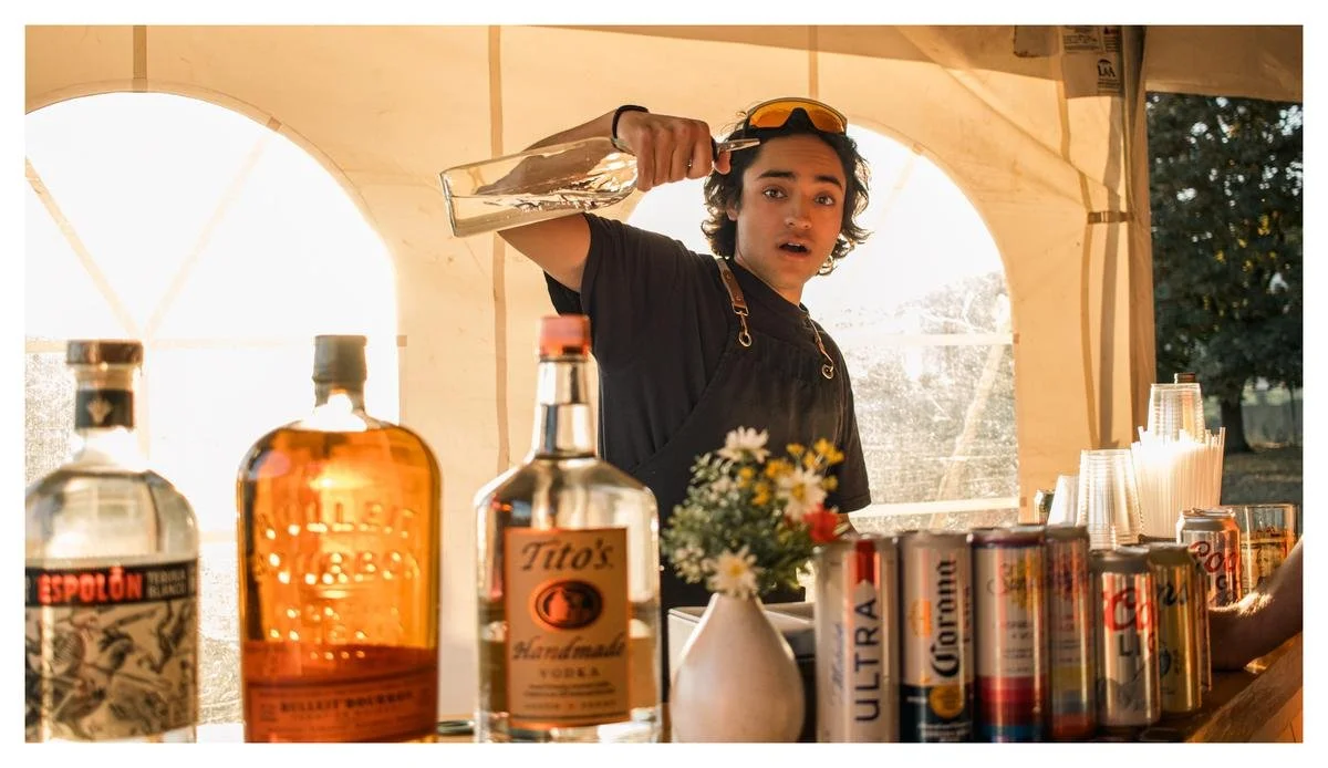 A young man with curly hair and sunglasses on his head stands behind a bar with bottles of alcohol and cans of beer, holding a large glass bottle with water or ice. He is wearing a black shirt and a black apron, inside a tent or outdoor bar setup, wi