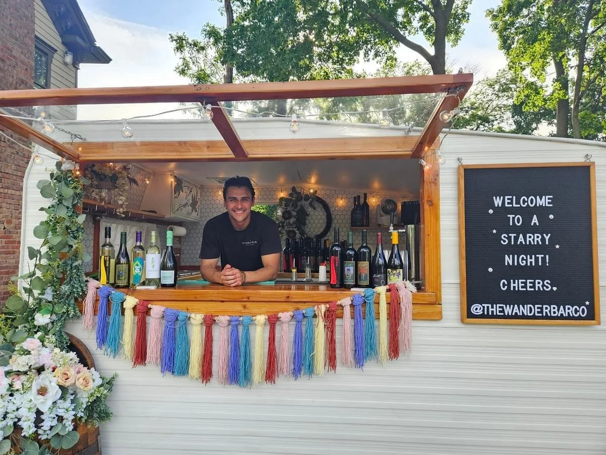 A smiling man standing behind a small outdoor bar in a white mobile trailer decorated with a rainbow-colored tassel garland, bottles of wine, and flowers. A sign on the side reads 'Welcome to a starry night! Cheers. @THEWANDERBARCO'. There are trees