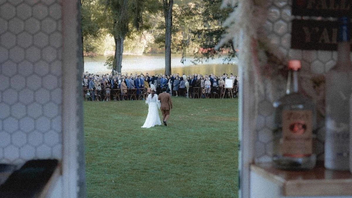 Bride and groom walking on grass towards a large crowd of guests near a lake, viewed from inside a building.