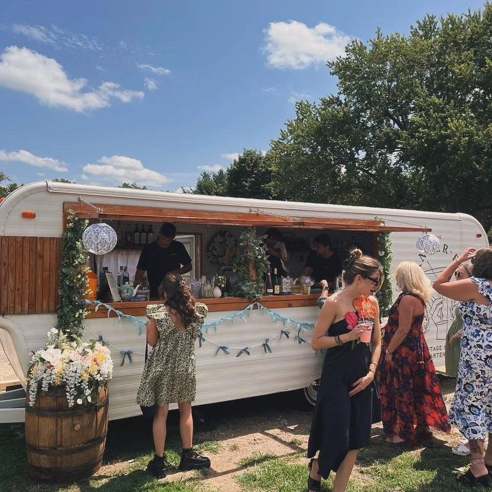 People at a mobile bar or food truck outdoors under a blue sky with white clouds, decorated with flowers and bunting.